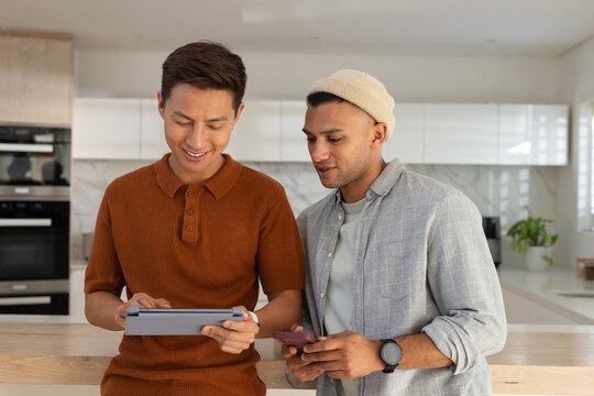 Asian and African American male friends standing in kitchen holding tablet and smartphone, leaning