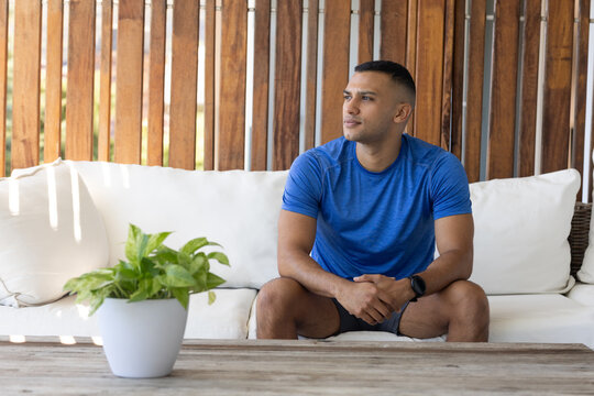 African American man sitting on sofa on patio, wearing blue tee, looking right, smartwatch, plant