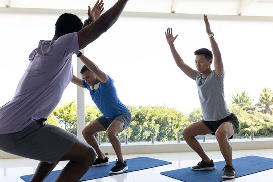 Diverse male group performing synchronized squats on blue exercise mats at bright glass studio