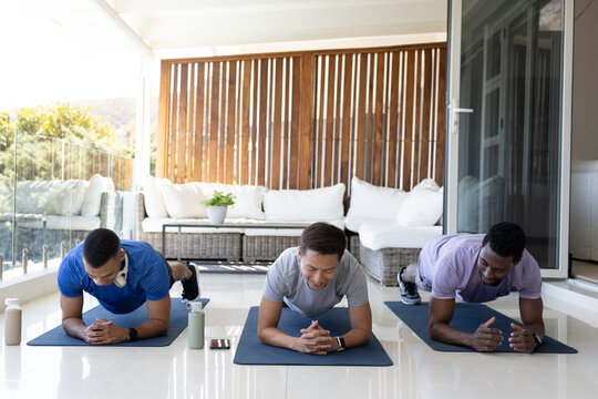Diverse male friends in sportswear holding forearm planks on veranda with dark mats, water bottles