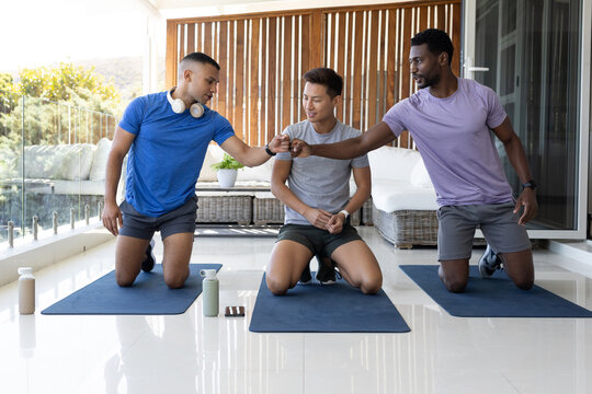 Diverse male friends kneeling, bumping fists in sportswear on patio with mats, water bottles, phone