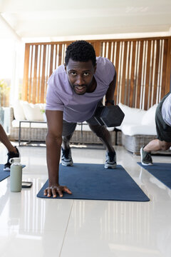 Diverse male workout group performing plank rows holding hex dumbbell on blue mats at home patio