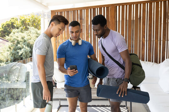 Diverse male friends leaning in and checking smartphone on covered balcony with yoga mats, bottles