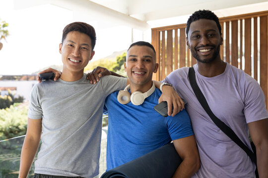 Diverse male friends standing on balcony, smiling at camera, holding yoga mat, headphones, phones