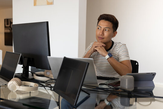 Asian man sitting at glass desk in home office using multiple monitors and headphones, copy space