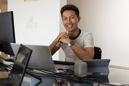 Asian man sitting in home office at glass desk smiling, clasping hands near chin, using laptop