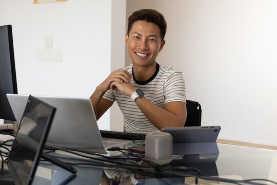 Asian man sitting in striped short-sleeve t-shirt at glass-top desk using laptop and monitor