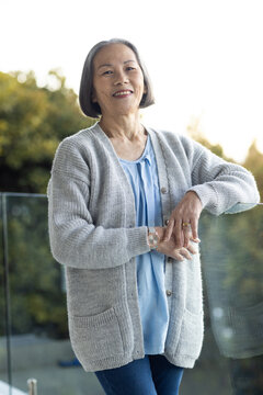 Senior Asian woman standing leaning on balcony glass railing in gray cardigan showing wristwatch