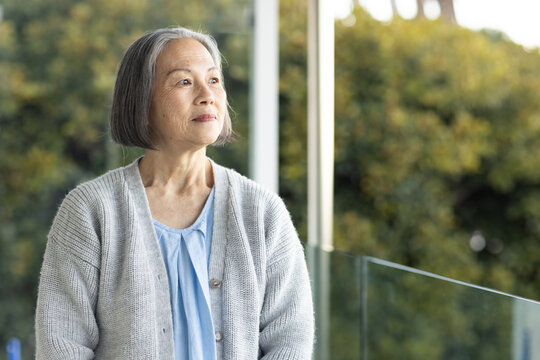 Senior Asian woman standing and gazing on home balcony by glass railing wearing grey cardigan