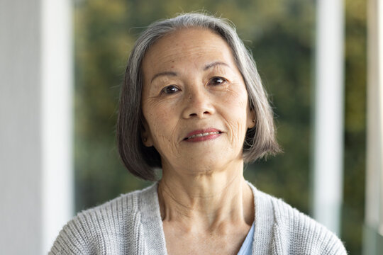 Senior Asian woman standing on porch wearing gray cardigan, blue top, stud earrings, garden behind