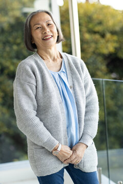 Senior Asian woman standing on balcony smiling, wearing grey cardigan, jeans, showing watch, ring