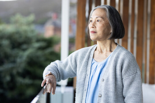 Senior Asian woman standing on balcony leaning on metal railing, wearing grey cardigan, wristwatch