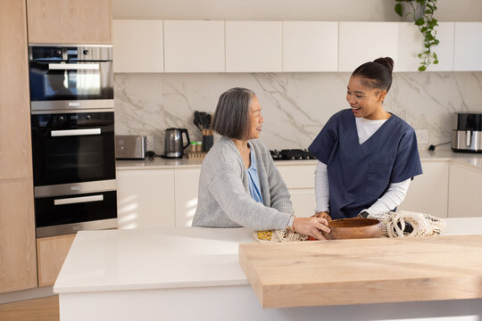 African American caregiver in scrubs and Senior Asian chatting at kitchen island, holding wood bowl