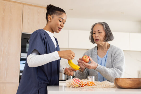 Two women, Senior Asian and African American caregiver in scrubs sorting fruit at kitchen island