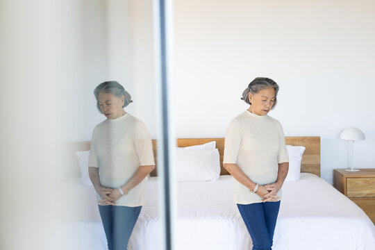 Senior Asian woman standing, clasping hands, wearing beige top, jeans in bedroom by mirror and bed