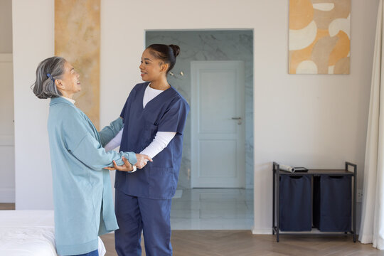 Female African American caregiver in navy scrubs helping Asian senior holding hands in room by bed