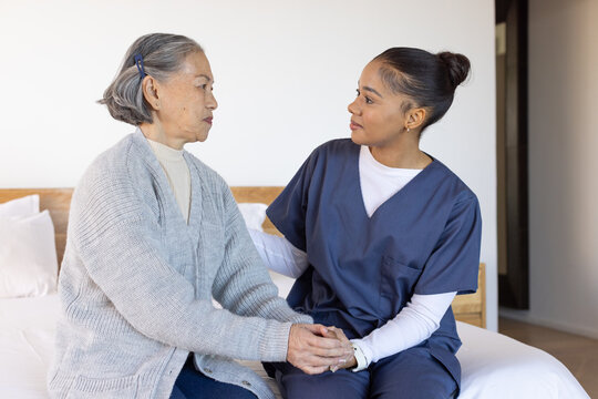 Senior Asian woman sitting on bed in bedroom holding hands with female caregiver in navy scrubs