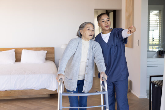 Senior Asian woman using walker while African American caregiver in navy scrubs pointing in bedroom