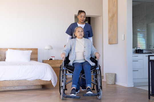 Asian senior sitting in wheelchair, African-American nurse in scrubs pushing in bedroom with meds