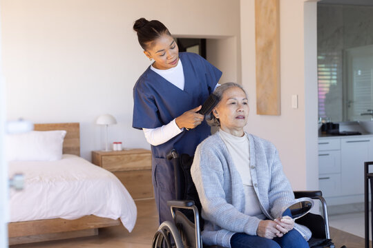 Senior Asian woman in wheelchair holding mirror, African American aide in scrubs grooming with comb
