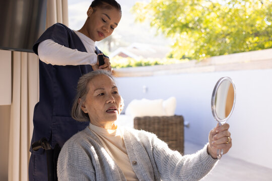 Senior Asian woman sitting on patio holding mirror while caregiver in scrubs using comb on hair