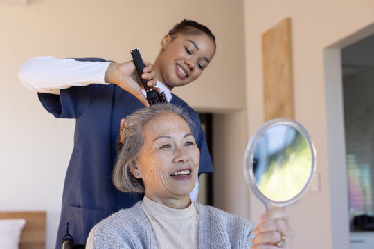 Diverse female stylist in navy smock brushing senior Asian client holding mirror and brush at home