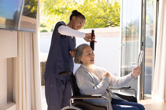 Asian senior woman in wheelchair African-American aide in scrubs using comb holding mirror at home