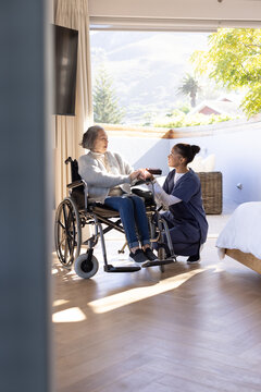 Senior Asian woman and African American nurse kneeling by wheelchair offering remote at bedside