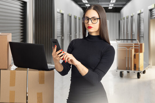 Self storage management, Female manager uses smartphone and laptop to coordinate inventory and customer orders in storage facility corridor.