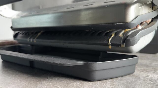 Excess fat and juice dripping from modern electric grill into plastic drip tray during meat cooking process in home kitchen. Healthy cooking and appliance concept