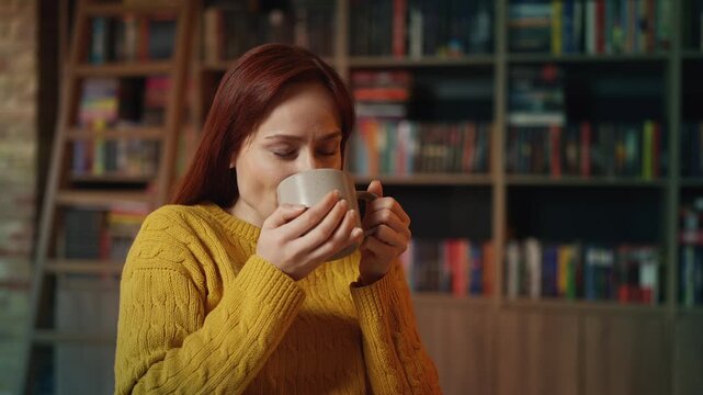 Positive red-haired female student enjoying warm tea during bookshop study break