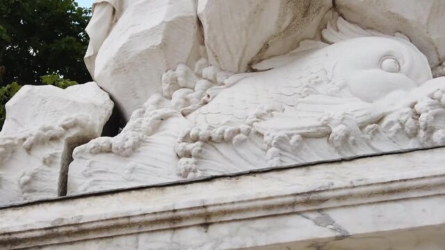 A magnificent detail of a white marble statue in a classical style (Baroque Classicism). The camera begins by focusing on the marble-textured base, then moves upward past carved motifs of water waves,