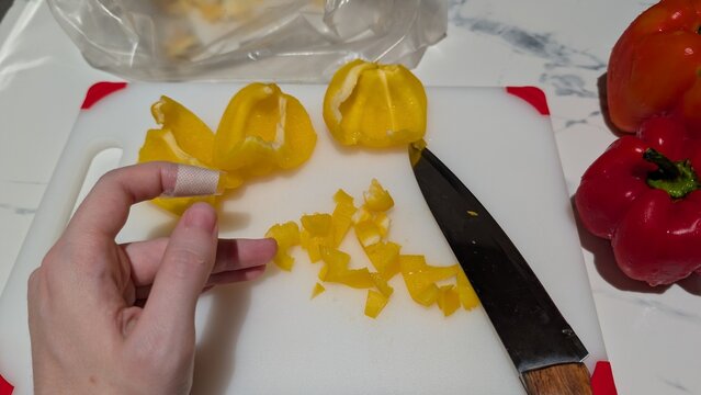 First aid for minor cut on finger while cooking. Left hand with bandaged index finger after knife injury in the kitchen. Cutting yellow bell pepper on plastic board background. 