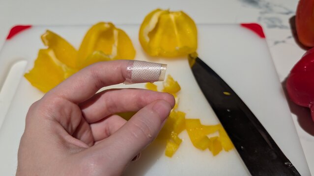 First aid for minor cut on finger while cooking. Left hand with bandaged index finger after knife injury in the kitchen. Cutting yellow bell pepper on plastic board background. 