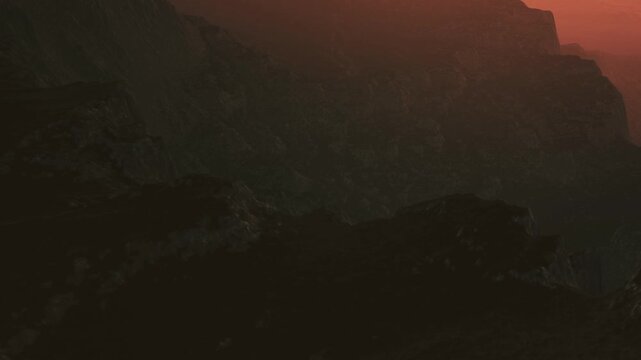 The scene shows rugged mountain terrain with varying shadows as the sun sets in the background. The colors shift with the light creating a dramatic view of the landscape.