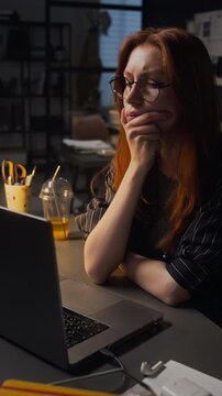Vertical video of young adult Caucasian woman with glasses typing on laptop, pausing to think, and resuming work late at night. Footage represents corporate overtime or freelance projects