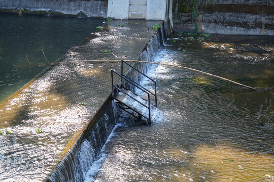 A black metal staircase submerged in water at a small river dam with flowing rapids.