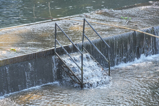 A black metal staircase submerged in water at a small river dam with flowing rapids.