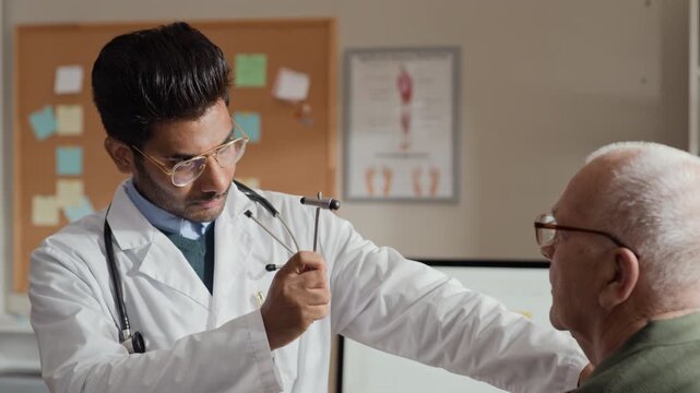 Young adult South Asian male doctor testing senior Caucasian male patient reflexes with neurological hammer. Perfect for geriatric healthcare marketing and medical clinic promotional videos
