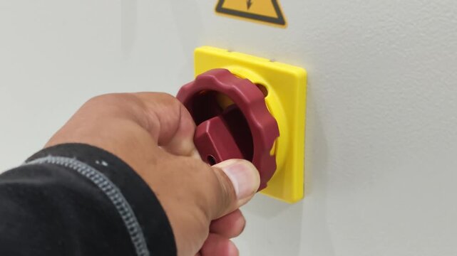 A close-up view of a technician's hand turning a red rotary power switch knob mounted on a yellow base on an industrial electrical control panel