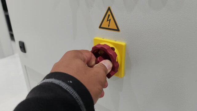 A close-up view of a technician's hand turning a red rotary power switch knob mounted on a yellow base on an industrial electrical control panel