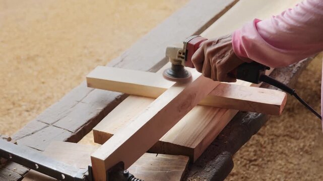 Carpenter's hands using an electric orbital sander to smooth a wooden plank in a woodworking workshop, part of the furniture making and finishing process.