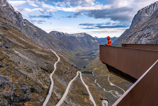 Tourist in orange jacket standing on Trollstigen viewing platform looking at dramatic winding mountain road in valley, Norway