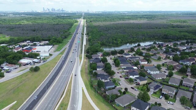 S. Buckner Boulevard runs parallel to electric rail line dense forest flanking suburban cul de sacs. Multi lane highway corridor stretches toward distant downtown skyline, suburbs Dallas, Texas