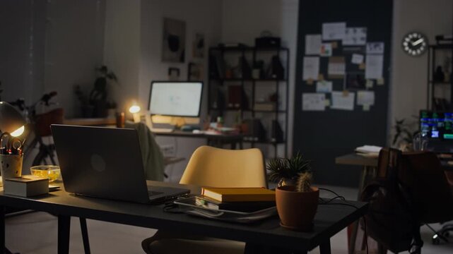 Panning shot of laptop computer sitting open on empty desk in creative office at night, representing overtime work. Ideal for corporate videos about business deadlines or remote work flexibility