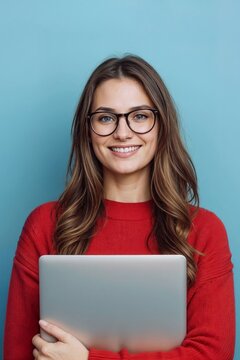 Portrait of young student woman smiles warmly while holding laptop on blue background. She wears red sweater and round glasses