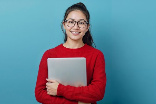 Young woman smiles warmly, holding laptop. Red sweater contrasts with soft blue background