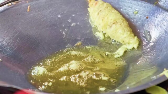 The tantalizing process of a rolled culinary treat deep-frying in shimmering hot oil within a wide wok, creating a crispy exterior for a popular street snack