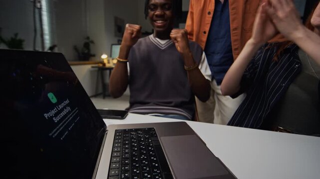 Diverse young adult colleagues celebrating successful project launch on laptop screen in office at night. Ideal for corporate teamwork, startup success, and software release marketing campaigns