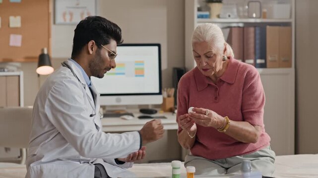 South Asian male doctor explaining prescription medication to senior Caucasian female patient in clinic. Footage ideal for healthcare marketing, geriatric care, and medical insurance advertising